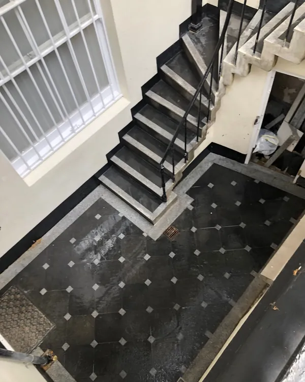 Looking down into a town-house lightwell: dark stone floor tiles with small pale squares where their corners meet, a grey stone border, and steps with dark treads and pale edges